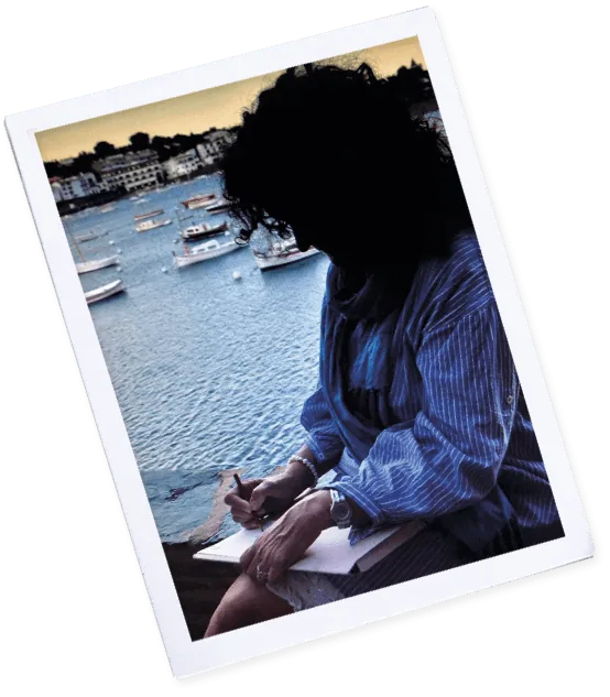 Polaroid de Laura dibujando junto al mar, con barcas al fondo